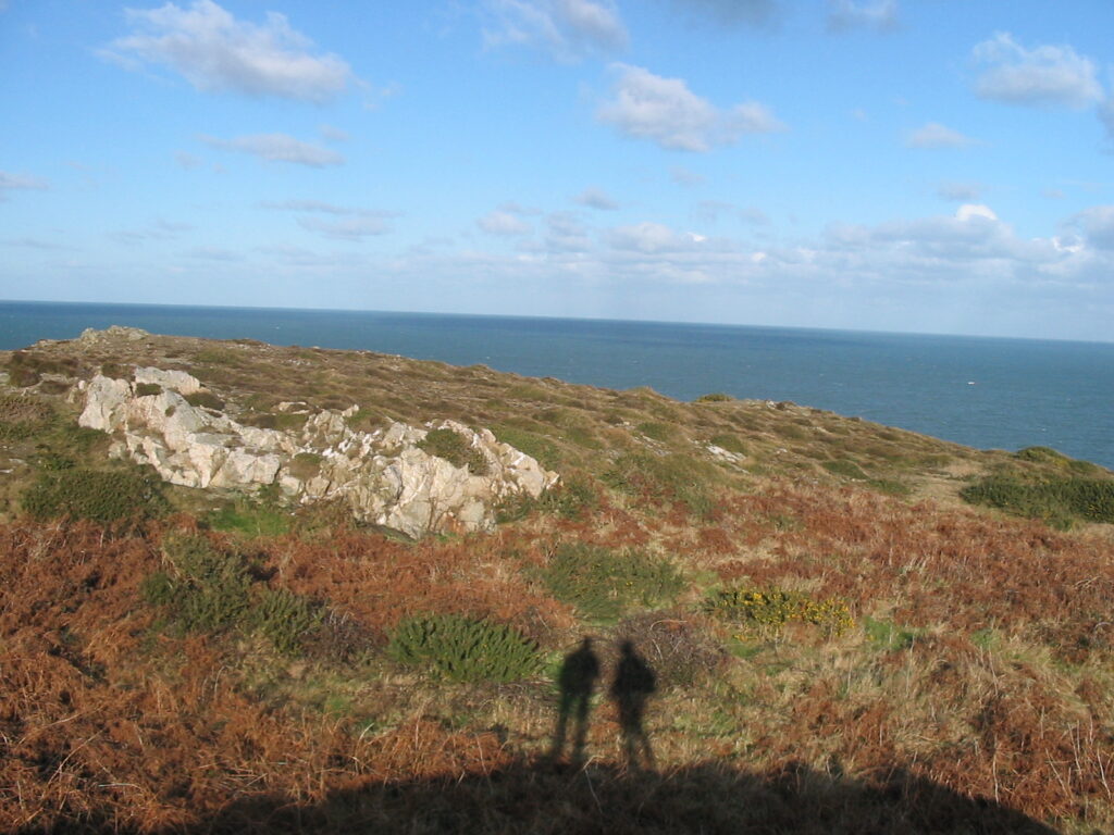 Foto einer kargen Landschaft vorm Meer. Im Vordergrund sieht man den Schatten eines Hügels, darauf der Schatten zweier Menschen.