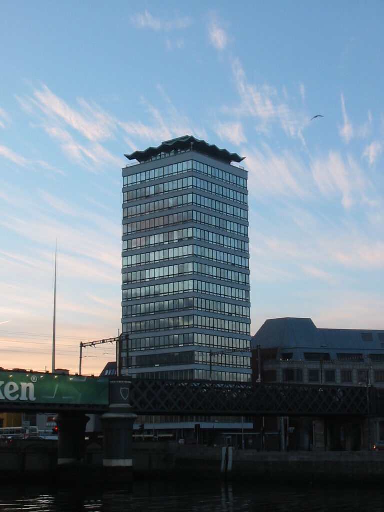 Ein 16stöckiges Hochhaus am Ufer eines Flusses. In den Fenstern spiegeln sich z. T. die Wolken.