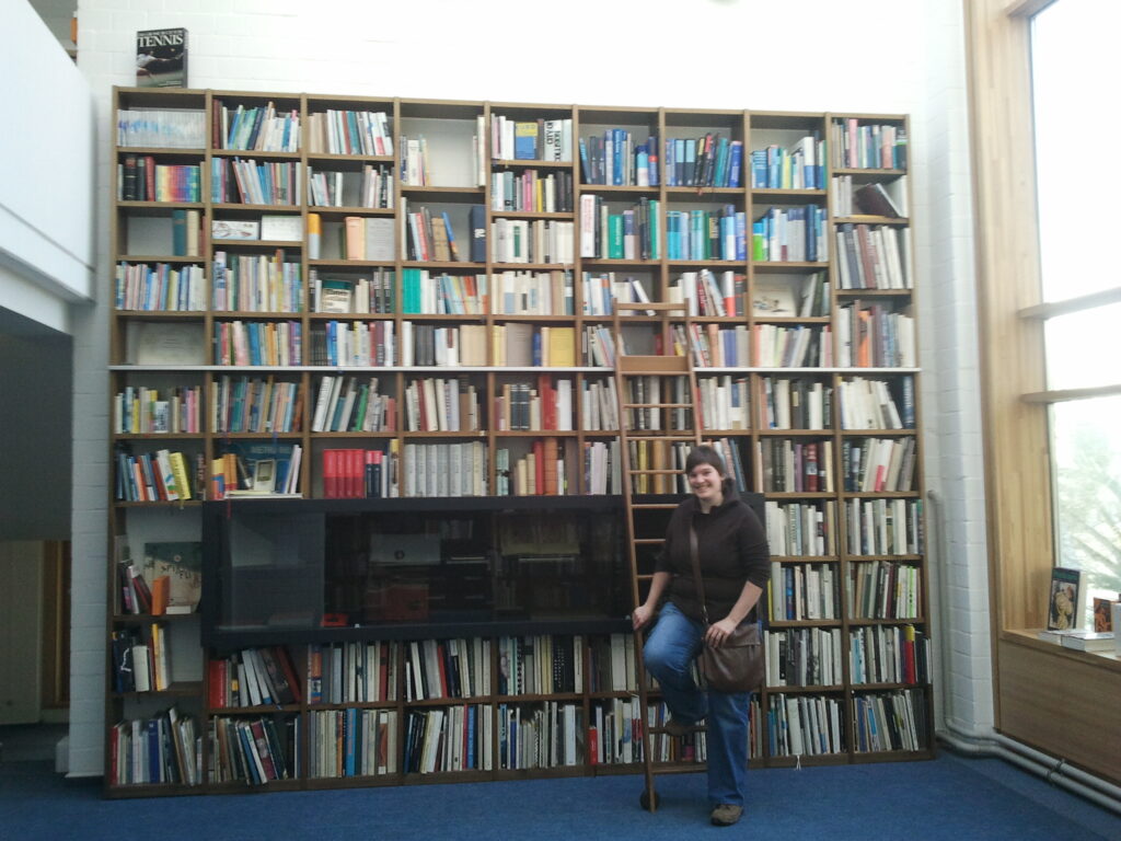 Foto. Carolin posiert vor einem riesigen Regal, das mit Büchern gefüllt ist.